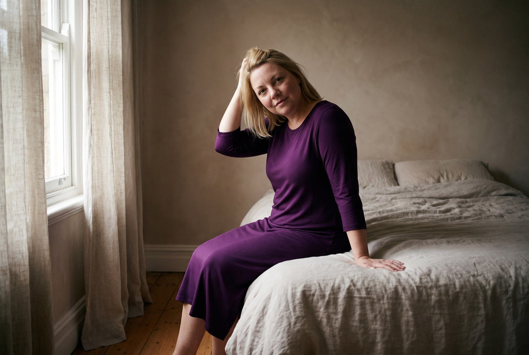 A woman in a Cool-jams NightCool nightgown in merlot, sitting on the edge of an unmade bed in early morning light.