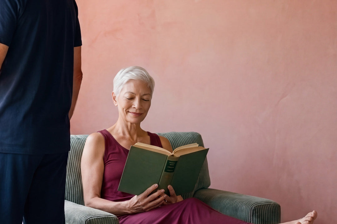 Woman reading in an armchair in a merlot NightCool long tank gown while a man in a navy NightCool tee and pants looks out the window