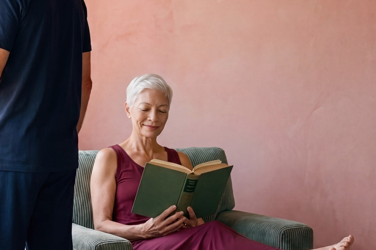 Woman reading in an armchair in a merlot NightCool long tank gown while a man in a navy NightCool tee and pants looks out the window