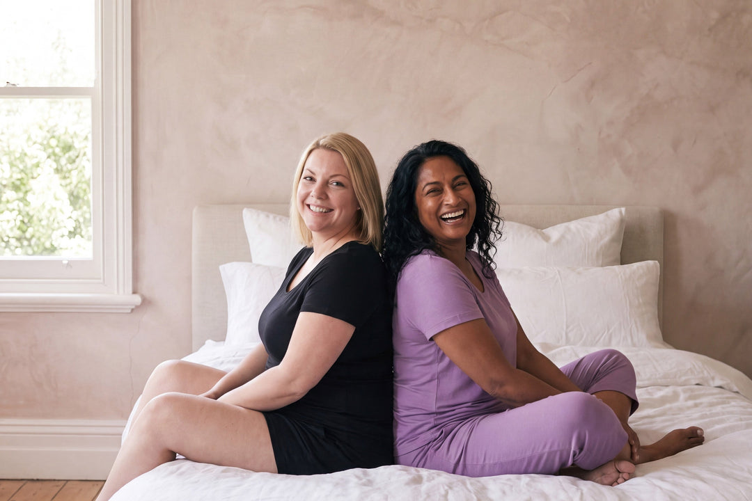 Two smiling women sitting back to back on a bed, one in a black NightCool scoop t-shirt and shorts, one in a violet scoop t-shirt and capri pants