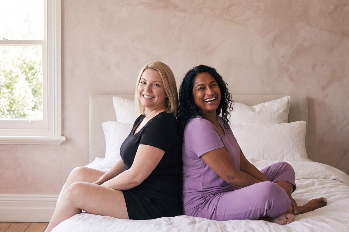 Two smiling women sitting back to back on a bed, one in a black NightCool scoop t-shirt and shorts, one in a violet scoop t-shirt and capri pants