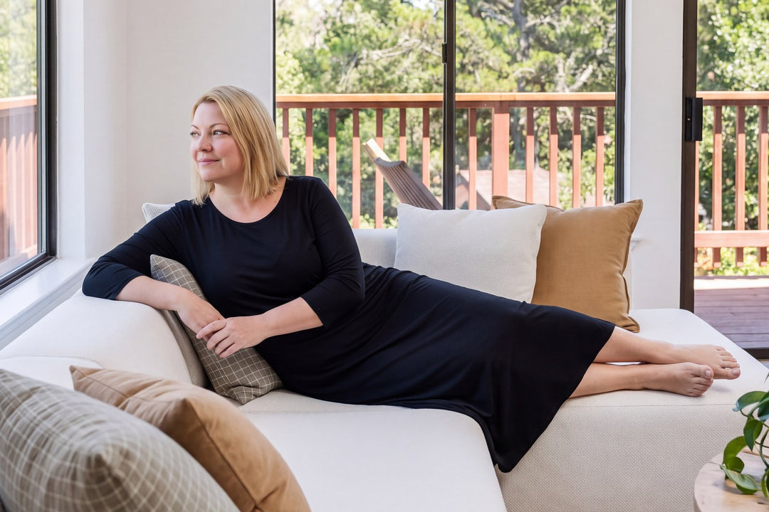Woman reclining on a white sofa by tall windows in a NightCool long scoop neck nightgown with 3/4 sleeves in Black, the sleeves ending at the forearm