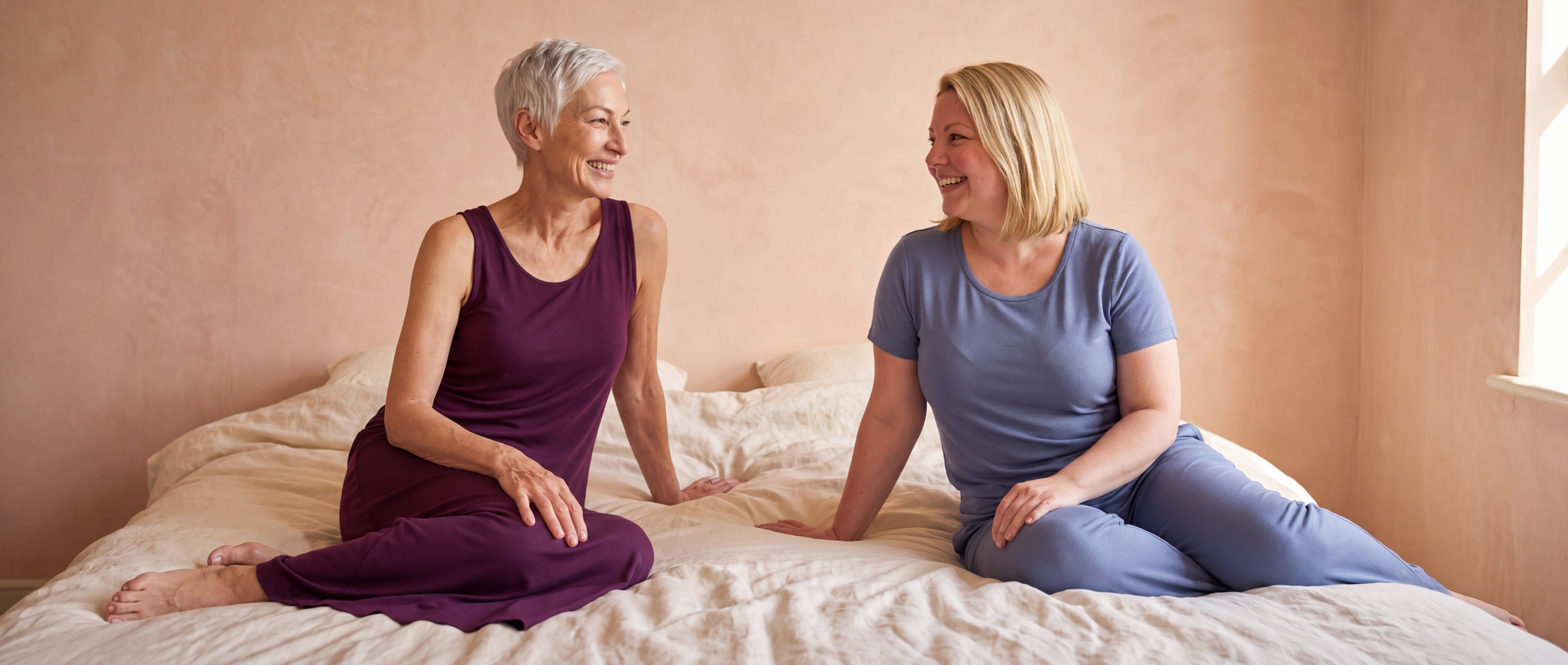 Two women laughing together on a bed in a pale coral plaster bedroom, one in a Merlot NightCool Long Tank Gown and one in a Dusk Blue NightCool scoop t-shirt and pants